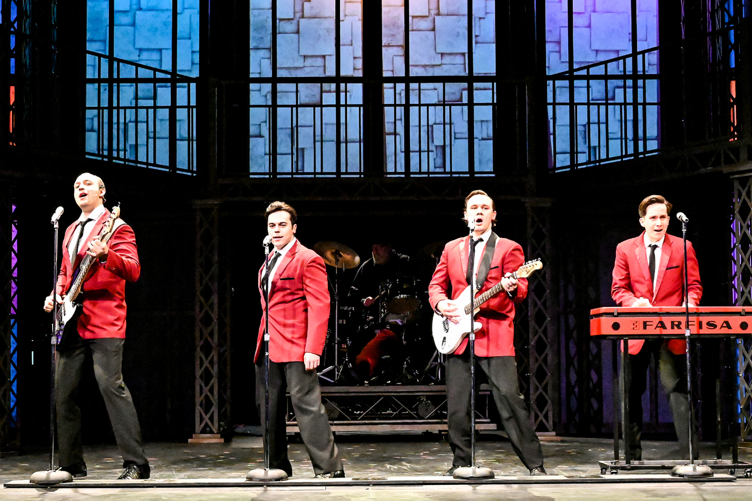 Production photo of four actors standing in a row, wearing red blazers, singing with microphones. The actors are depicting the band, the Four Seasons. 