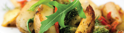 A close-up shot of a plate of roasted vegetables, including potatoes, broccoli, and red peppers. A large, bright green arugula leaf is prominently placed in the center of the dish. The vegetables are lightly browned and appear to be seasoned, suggest