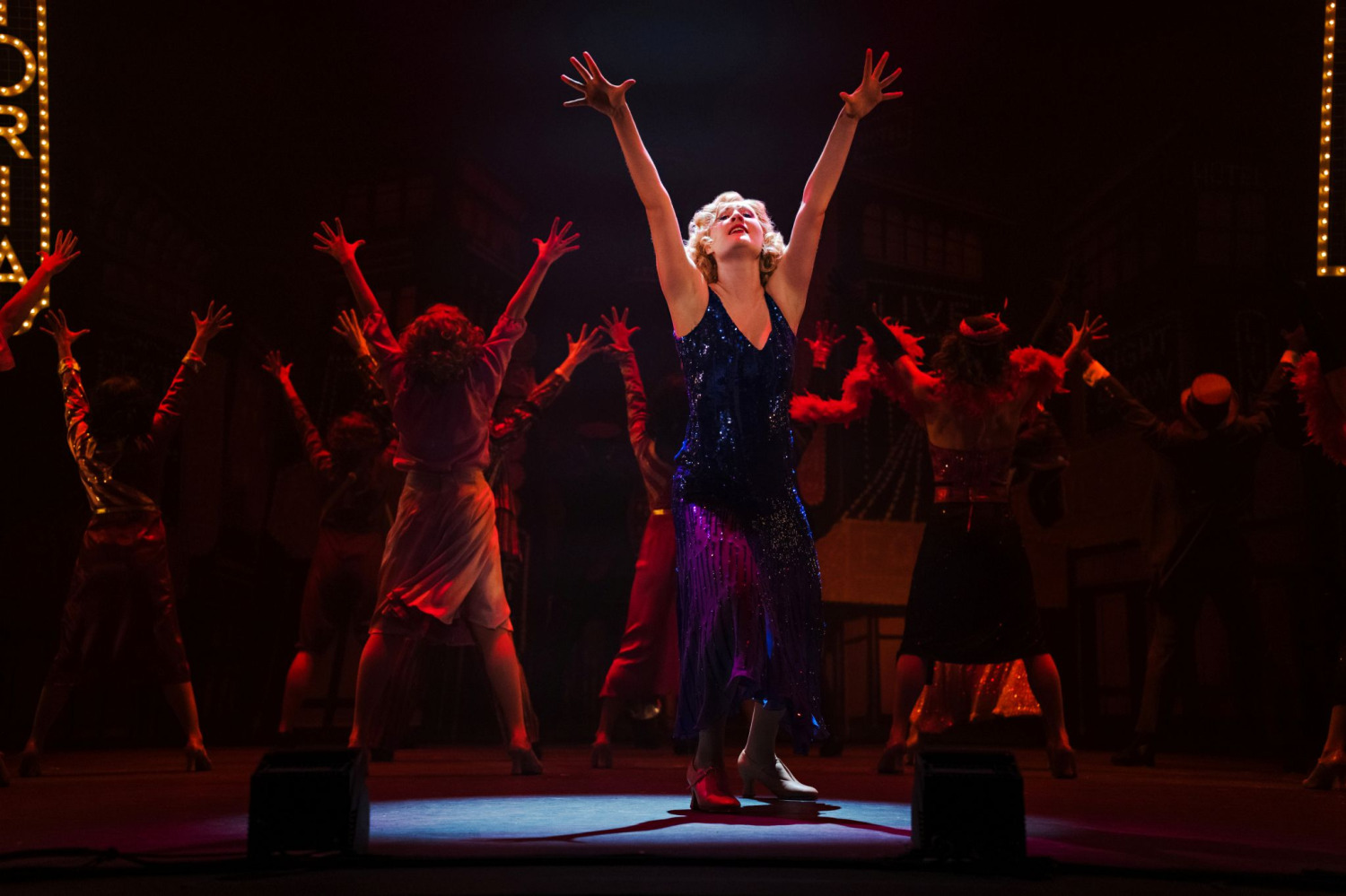 A performer in a sparkling blue dress stands in a spotlight during a dramatic musical number from 42nd Street. Bathed in rich red lighting, they reach both arms upward while ensemble members pose behind them with matching gestures. Marquee-style Broadway signs and cityscape scenery frame the powerful performance.