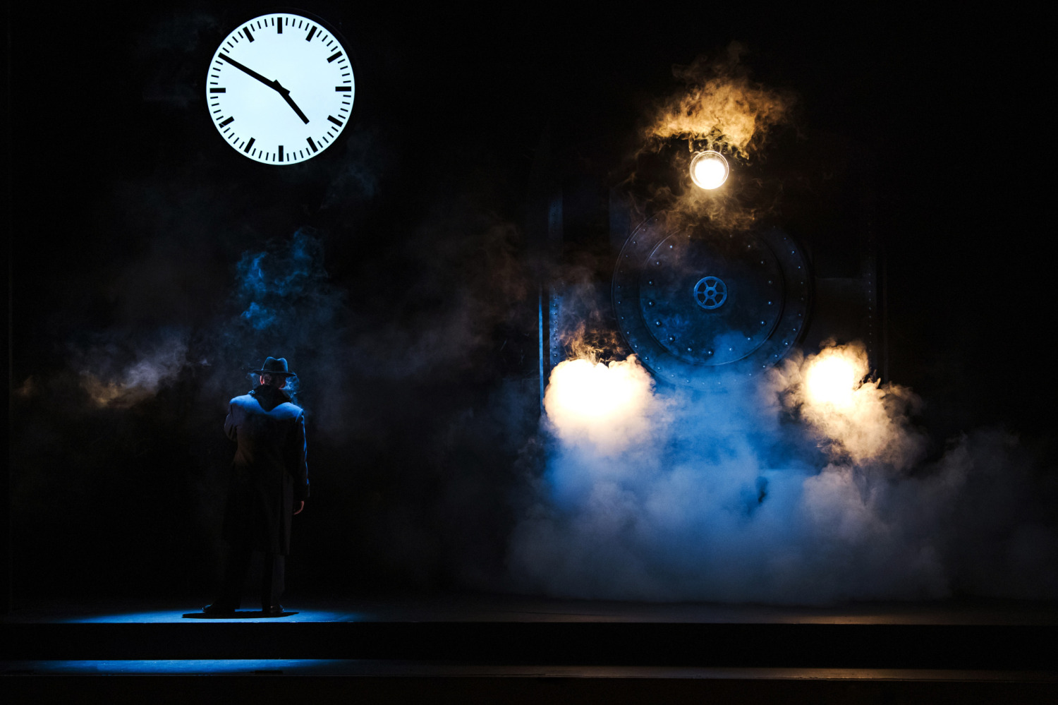 A lone figure stands on a dark stage facing a steam locomotive surrounded by dramatic blue and amber light, thick smoke, and a large illuminated clock overhead.