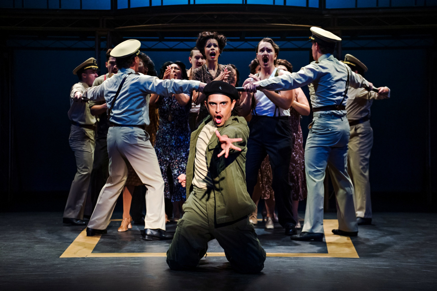 A dynamic moment as a character in military fatigues and a beret kneels centre stage, reaching forward with intensity. Behind him, uniformed officers form a barrier with outstretched arms, attempting to hold back a distressed crowd of citizens who shout and reach forward. The scene is charged with emotion and urgency, highlighted by stark lighting and a bold blue backdrop.
