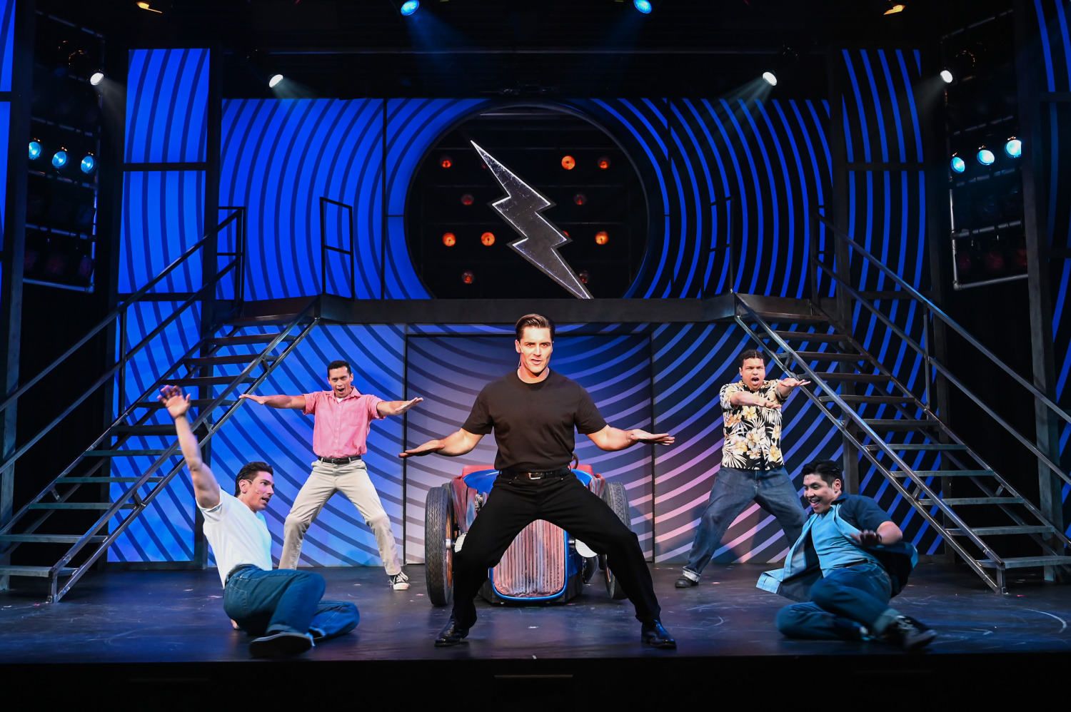 A dynamic performance scene from Grease features six male performers mid-dance in front of a stylized, blue-striped set with a large lightning bolt emblem. One performer in a black t-shirt stands centre stage in a wide stance, arms outstretched, while two others slide across the floor dramatically. The remaining performers strike bold poses around a vintage red and blue car prop, creating a visually striking tableau full of energy and retro flair.