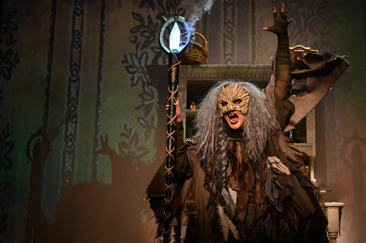 In Drayton Entertainment’s production of Into the Woods at St. Jacobs Country Playhouse, a performer portraying the Witch stands centre stage in a ragged, layered brown costume with long grey hair and a textured, bark-like mask. They grip a tall black staff topped with a glowing blue crystal as smoke curls from it, while one arm reaches dramatically overhead. Dark floral and woodland projections cover the set behind them, creating an eerie, magical atmosphere.