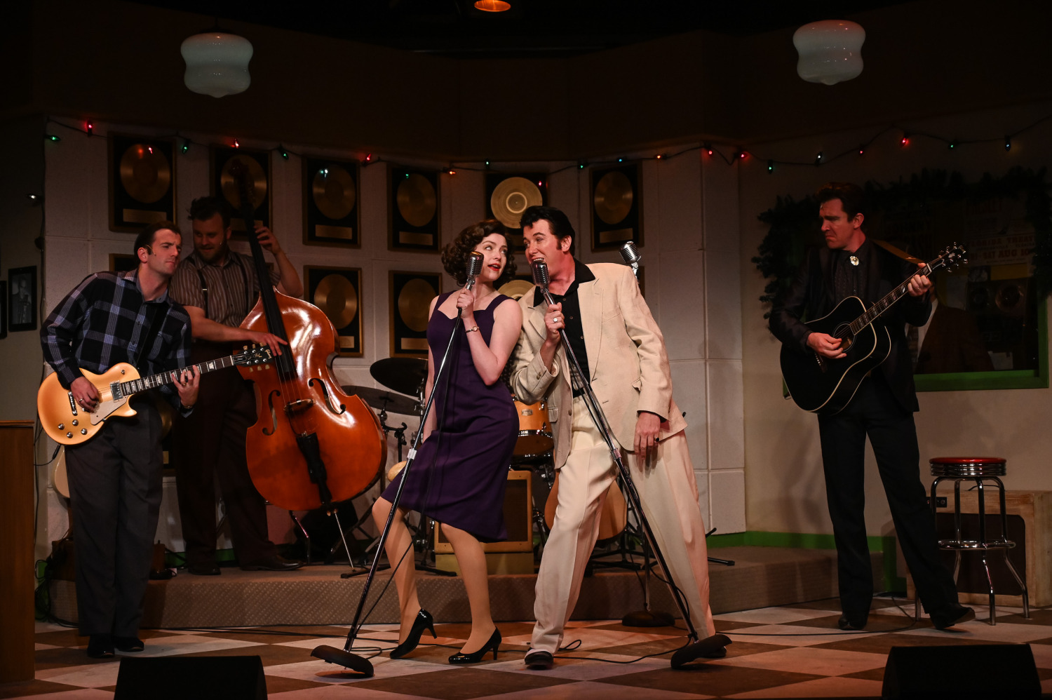 A lively musical number from Million Dollar Quartet, set in a vintage recording studio. At centre stage, a performer in a cream-coloured suit and a performer in a fitted purple dress sing into retro microphones, leaning in playfully toward each other. Behind them, a guitarist in a plaid shirt, an upright bassist, and a man in a black suit with a black guitar round out the band. The set is decorated with gold records, holiday string lights, and recording studio details, evoking a mid-century rock-and-roll atmosphere.