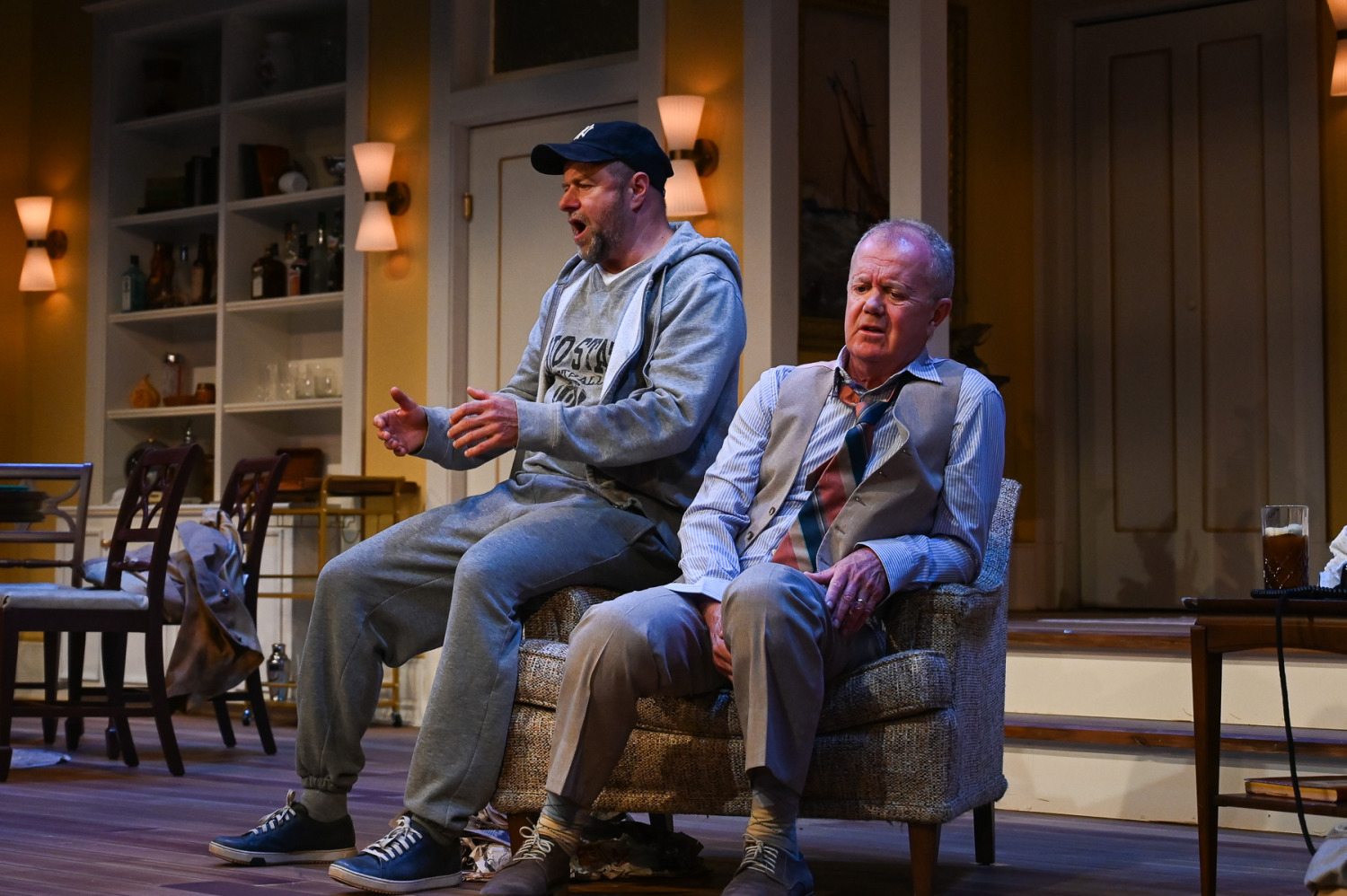 Two performers share a comic moment in the living room. One, dressed in grey sweats and a baseball cap, perches on the arm of a chair with his mouth open and hands extended, while the other slumps in the chair wearing dress pants, a vest, and a loosened tie.
