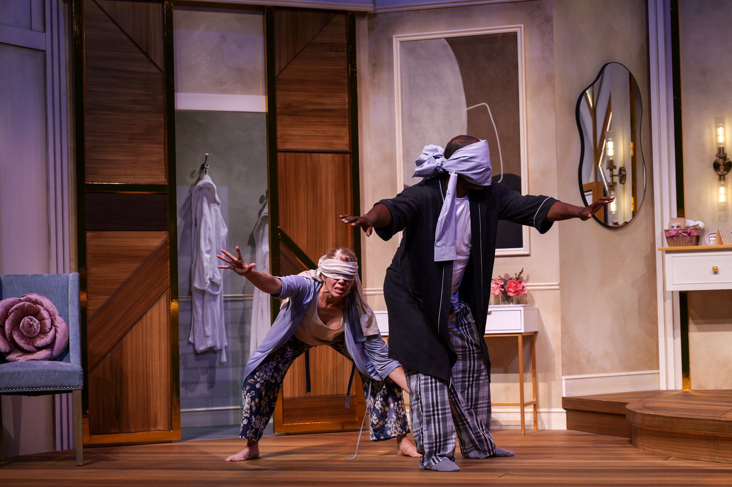 Two blindfolded performers cautiously make their way across the bedroom set with their arms stretched out in front of them. One bends low while reaching forward, while the other stands behind with a piece of fabric tied dramatically around their head and eyes.