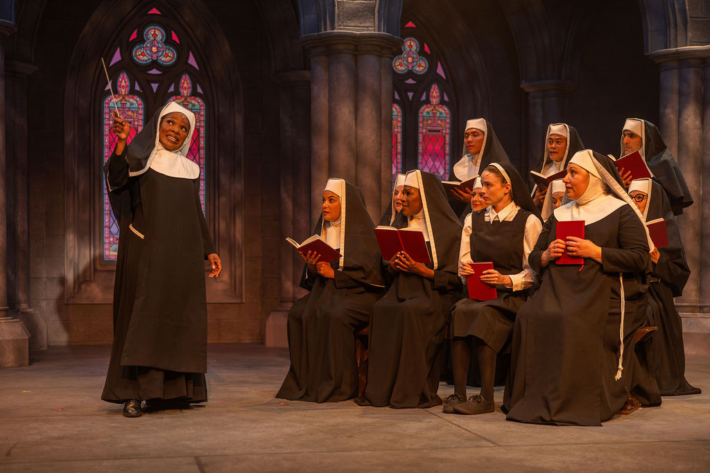 Promotional image for Sister Act at King’s Wharf Theatre, featuring bold red title lettering against a radiant blue background with sparkles and stained glass windows at the bottom.