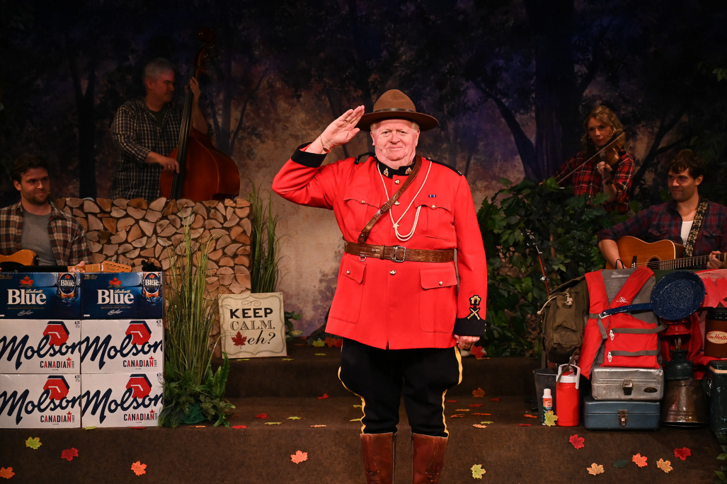 A comedic tableau unfolds in Sorry... I’m Not Sorry as a performer dressed in the iconic red uniform of a Royal Canadian Mounted Police officer stands centre stage delivering a crisp salute. Wearing the traditional red serge jacket, wide-brimmed hat, riding boots, and ceremonial belt, the performer strikes a proud pose while musicians playing guitar, fiddle, and upright bass accompany the scene from behind. The woodland-themed set is filled with camping gear, coolers, autumn leaves, stacked cases of Molson Canadian and Labatt Blue, and a sign reading “Keep Calm, eh?”, creating a playful and distinctly Canadian atmosphere that celebrates one of the country’s most recognizable national symbols.