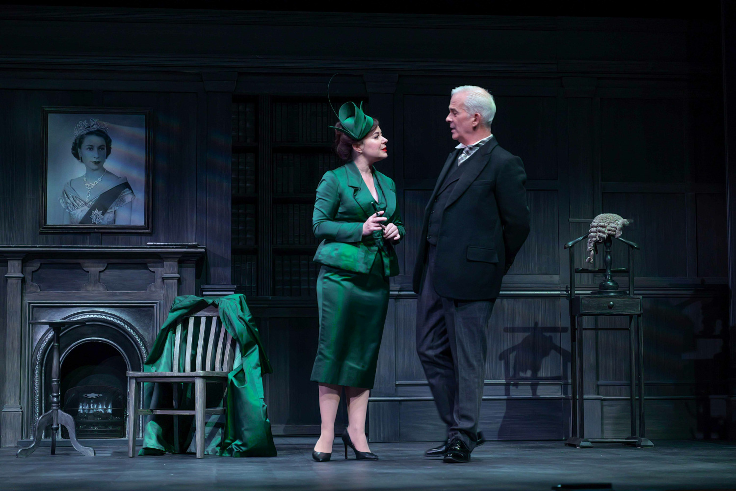 In this scene from Agatha Christie’s Witness for the Prosecution a woman in an elegant emerald-green suit and matching hat stands speaking to a man in a dark suit within a dimly lit, wood-panelled study. Behind them, a portrait of Queen Elizabeth II hangs above a fireplace, and a barrister’s wig rests on a stand nearby. The woman’s confident posture and the man’s attentive stance suggest a charged, secretive exchange in this tense British courtroom drama.