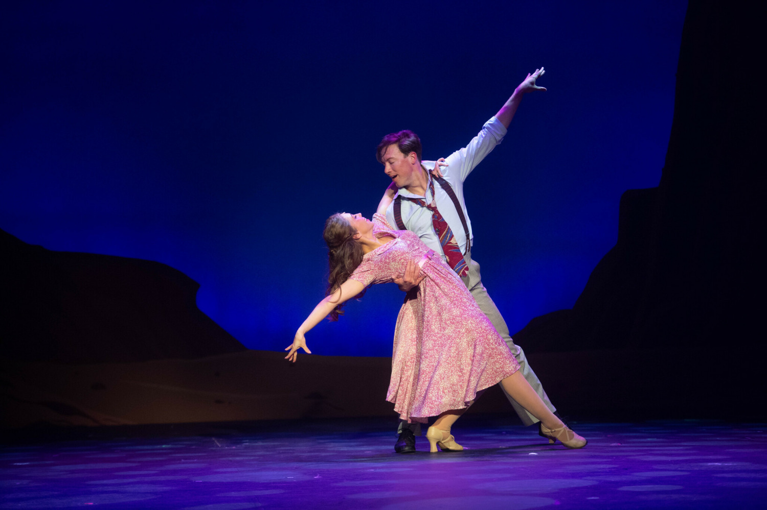 In this image from Crazy For You, a romantic dance moment on stage featuring two performers. The dancer in a pink floral dress is gracefully dipped by a partner in dress pants, a button-down shirt, suspenders, and a patterned tie. Their arms extend in elegant lines against a deep blue backdrop, with silhouetted desert hills framing the scene, evoking a dreamy, intimate atmosphere.