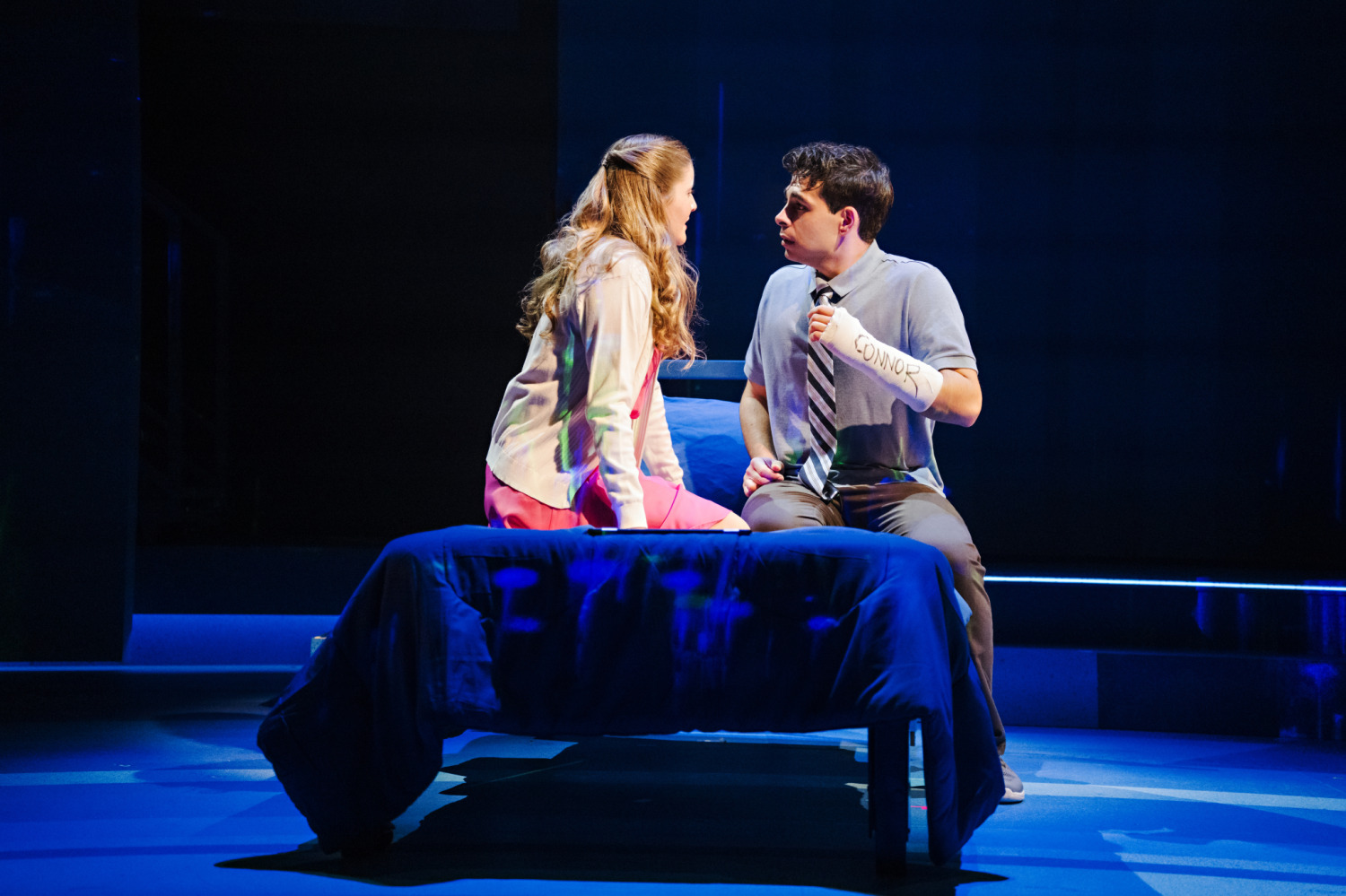 Two teenagers sit facing each other on a bed in Dear Evan Hansen. The boy, wearing a cast labelled “Connor,” leans forward anxiously while the girl in a pink skirt and light jacket listens intently, both framed in soft blue lighting that highlights the emotional intimacy of the moment.