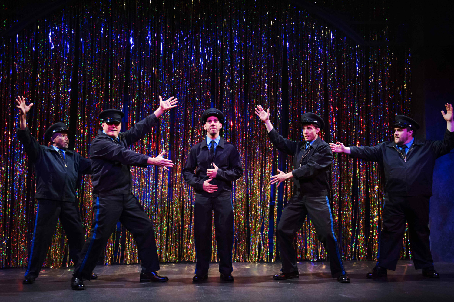 A comedic and theatrical moment from The Full Monty featuring five performers dressed as police officers posing dramatically in front of a shimmering, rainbow-coloured tinsel curtain. The central performer stands still with a serious expression while the others gesture flamboyantly toward him, striking exaggerated stage poses. The sparkling backdrop and playful staging suggest a humorous performance or talent show scene.