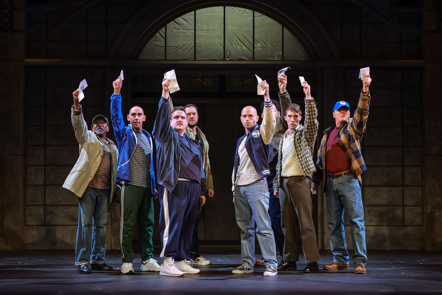 Seven men stand shoulder to shoulder on stage in The Full Monty at Huron Country Playhouse, each raising an envelope high above their heads in a powerful moment of solidarity. Dressed in everyday clothing ranging from work jackets and track suits to jeans and flannel shirts, the performers face the audience with determined expressions beneath warm theatrical lighting, framed by the entrance of an industrial-style building that reflects the show's working-class setting.
