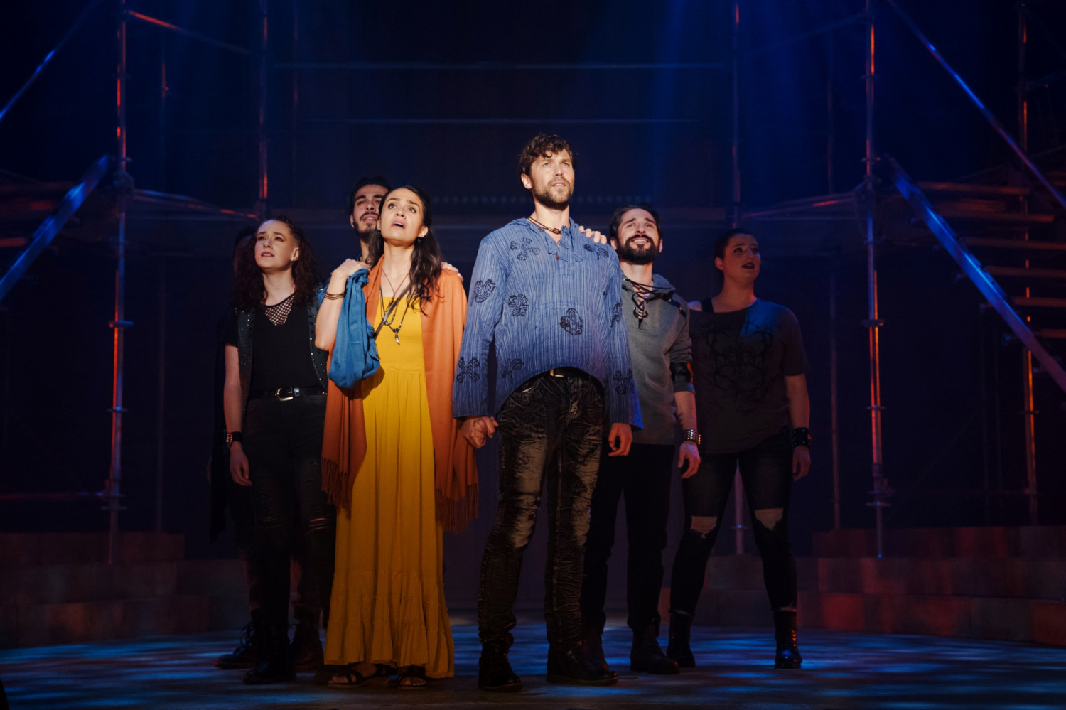 The promotional image for Jesus Christ Superstar features the title in bold, white letters against a sparkling, bokeh background. The backdrop transitions from brightly illuminating a cross outline in the center to shadow and smoke in the perimeter