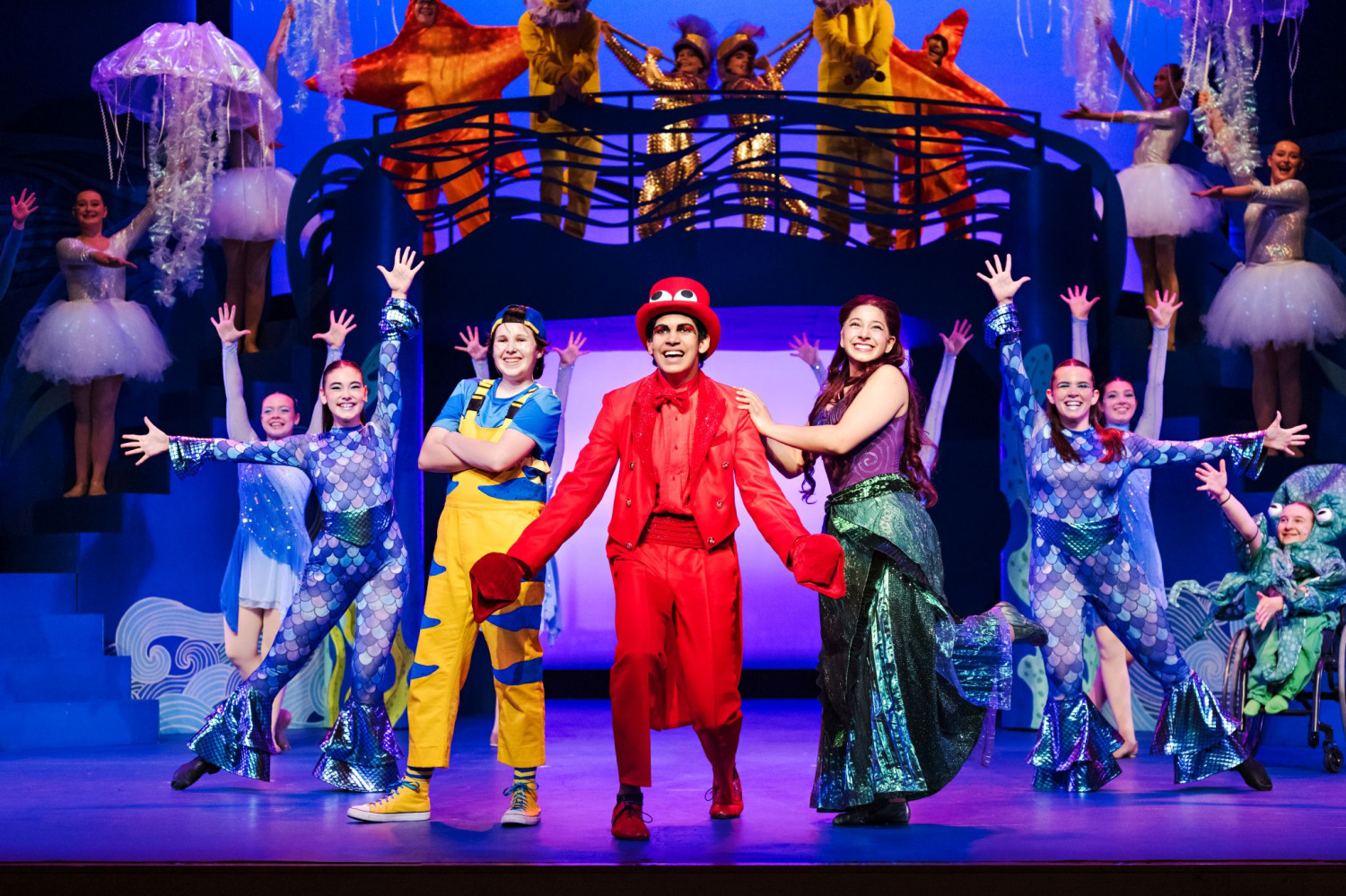 In a lively ensemble number from Drayton Entertainment’s Disney’s The Little Mermaid at Hamilton Family Theatre Cambridge, Ariel poses beside performers in bright red and blue-and-yellow costumes at centre stage while dancers in shimmering blue fish costumes extend their arms around them. Additional sea creatures and performers fill the raised platforms behind them beneath vivid blue and purple lighting, creating a colourful underwater spectacle.