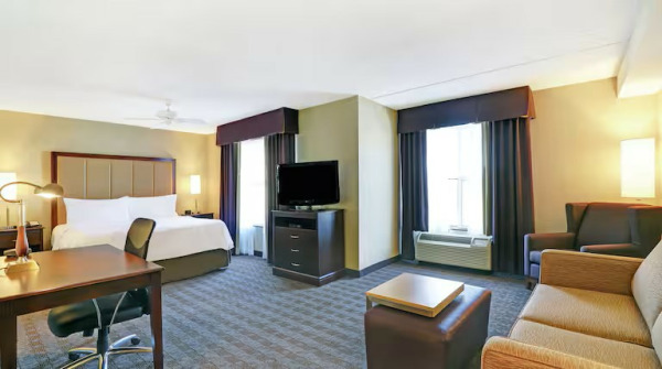 A spacious hotel suite with a modern and cozy design. The room features a large king-size bed with white linens, a wooden headboard, and bedside lamps. A work desk with an ergonomic chair and a desk lamp is positioned near the bed. The living area in