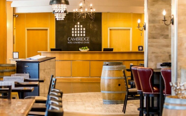 A warmly lit hotel lobby and restaurant area with a mix of modern and rustic decor. The reception desk in the background has a sign reading 'Cambridge Hotel & Conference Centre.' The seating area includes black leather chairs and wooden tables, with