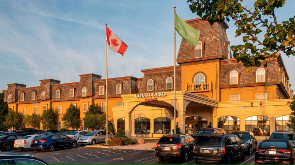 A daytime exterior shot of the Courtyard by Marriott Waterloo St. Jacobs hotel. The building is a multi-story structure with a yellow facade and a distinctive mansard roof with dormer windows. Two flagpoles stand in front, one flying the Canadian fla