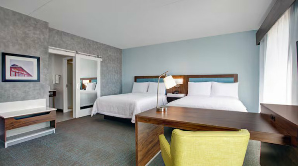 A bright and clean hotel room interior with two queen beds made with white linens. The room features a light blue accent wall behind the beds, a wooden desk with a yellow chair in the foreground, and a sliding barn door leading to the bathroom. A fra