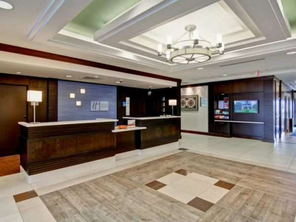 A well-lit hotel lobby featuring a dark wood reception desk with a white countertop. Behind the desk is a blue accent wall with the Holiday Inn logo. The lobby has light-colored tile flooring with a geometric pattern, and a decorative chandelier hang