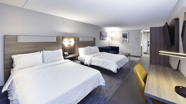 Modern hotel room featuring two queen beds with white linens, a wooden headboard with built-in lighting, a navy-blue loveseat, a flat-screen TV mounted on the wall, a wooden desk with a yellow chair, and contemporary artwork. The room has a neutral c