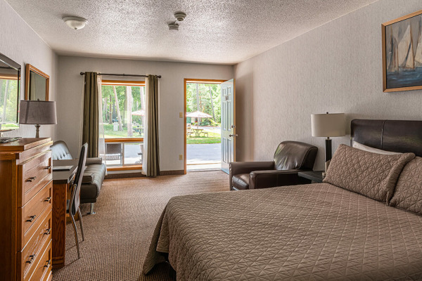 A modestly furnished motel room with a king-size bed featuring a quilted beige bedspread and matching pillows. A brown leather armchair sits beside the bed, and a wooden dresser with a mirror is on the left. A small sofa is placed near a window with
