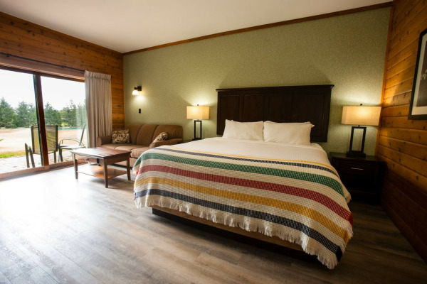 A cozy resort room with a large king-sized bed covered in a striped multicolour blanket. The room features wood-paneled walls, a small striped sofa with throw pillows, a wooden coffee table, and two bedside lamps. A sliding glass door opens to a pati