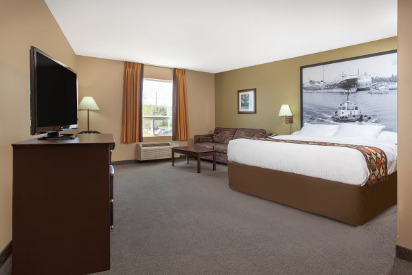 A clean, spacious hotel room featuring a comfortable king-sized bed, a cozy sitting area with a sofa and coffee table, a flat-screen TV, and warm neutral décor with natural light from the window.