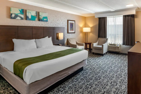 A spacious and elegantly styled hotel room featuring a comfortable king-sized bed, cozy sitting area with armchairs and a side table, soft ambient lighting, and large windows that fill the room with natural light.