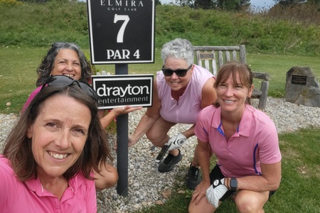 A Ladies foursome wearing pink golf shirts are posing around the marker for the 7th Hole at Elmira Golf Club which is hosted by Drayton Entertainment.