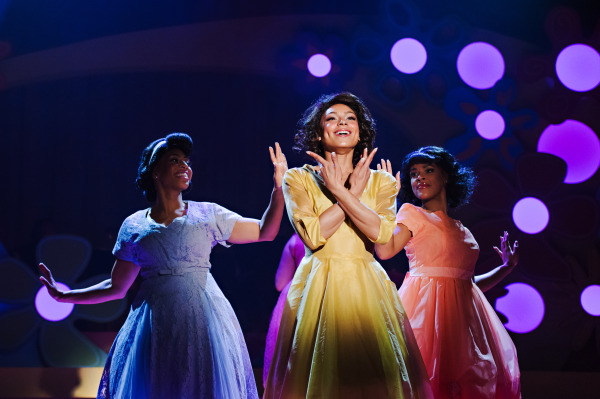 A production photo of three ladies in bright pastel dresses. the one in the middle fully lit by a spotlight