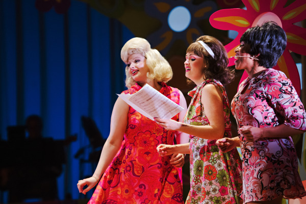 a production photo of 3 women dresses in busy flower and paisley print dresses and beehive hairstyles gather around sheet music as they sing 
