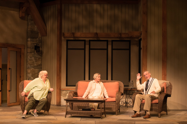 A scene from Doris and Ivy in the Home with three people sitting on a wicker sofa and chair, engaged in conversation, with a rustic indoor set in the background.
