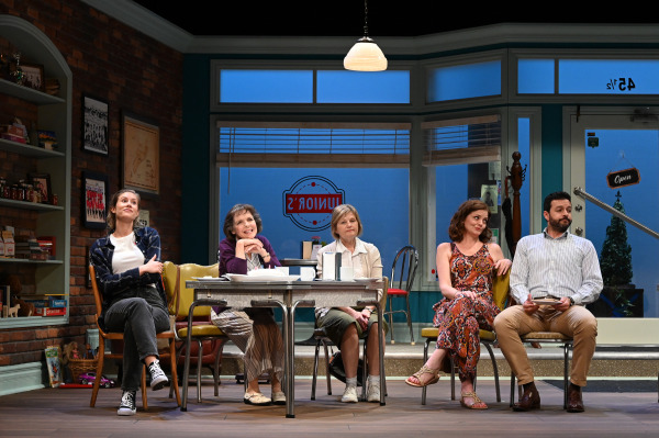 Five actors sitting at a diner table, engaging in conversation. The diner features a blue and white color scheme with a sign that reads 'Junior's'.
