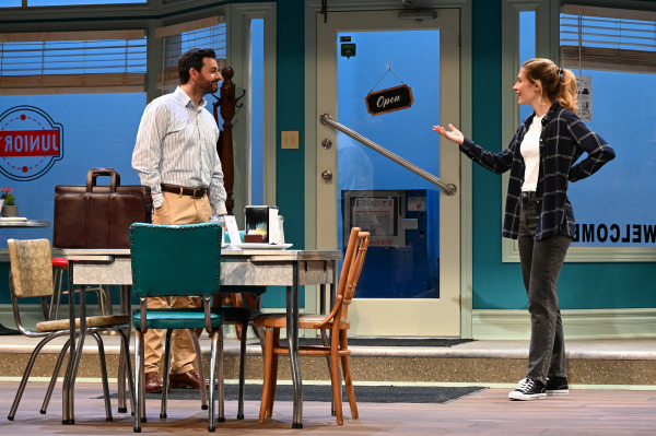 Two actors standing in a diner set, engaged in a conversation. One actor gestures with open arms while the other stands near a table. The diner features a blue and white color scheme with a sign that reads 'Open'