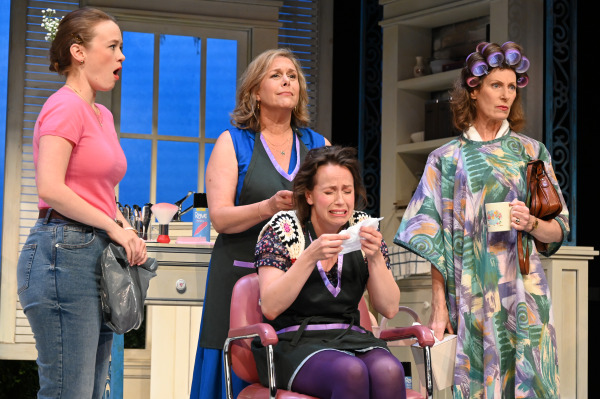 A dramatic scene from Steel Magnolias with four people in a beauty salon, one seated and visibly upset while the others stand around, each with varied expressions of concern and surprise.