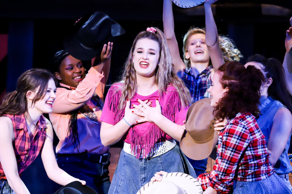 Production photo of Footloose showing a group of teenage girls holding cowboy hats and singing.