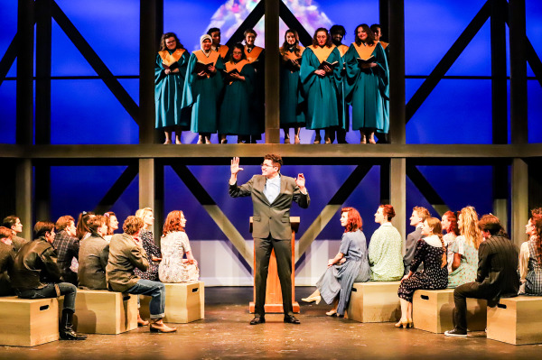 Production photo of Footloose showing the entire company of actors on stage, sitting in church pews and listening to a sermon.