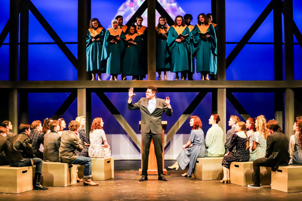 Production photo of Footloose showing the entire company of actors on stage, sitting in church pews and listening to a sermon.