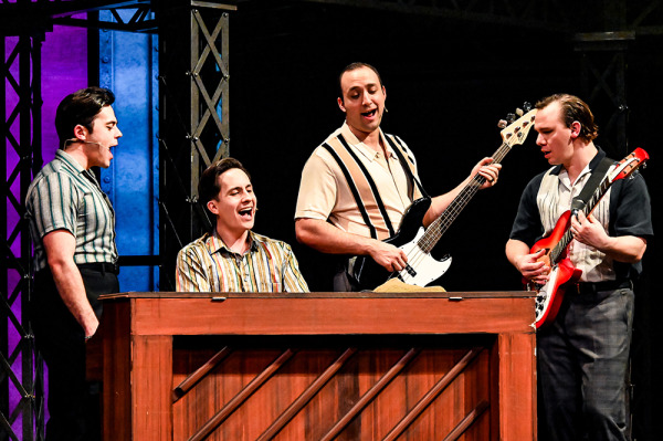 Four actors sitting around a piano singing. 