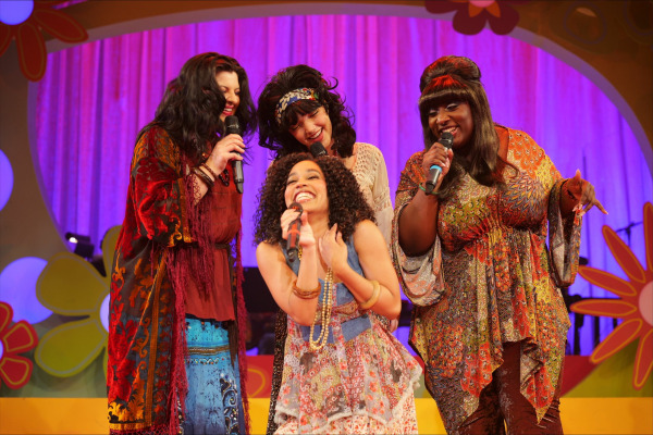 A lively scene from this musical celebration of the '60s and '70s features four performers standing close together mid-song, each holding a microphone and smiling brightly. They wear bold, patterned bohemian costumes with bell sleeves, layered skirts, and beaded accessories, evoking the free-spirited style of the era. The colourful set behind them is decorated with oversized daisies and bright purple curtains, enhancing the show's retro, flower-power aesthetic.