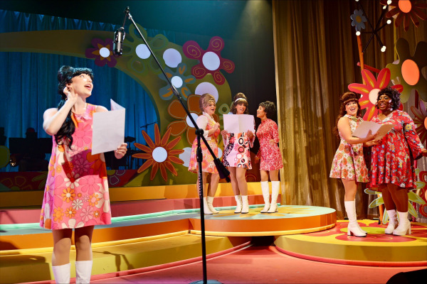 A vibrant scene shows seven performers mid-rehearsal, each holding sheet music and dressed in bold, brightly patterned mini dresses with go-go boots, channeling the energy of 1960s girl groups. One performer stands in front of a vintage microphone with hand to ear, while others cluster in groups of two or three, joyfully singing and laughing. The retro set features large flower cut-outs, swirling orange and pink platforms, and shimmering gold curtains, creating a playful, flower-power vibe.