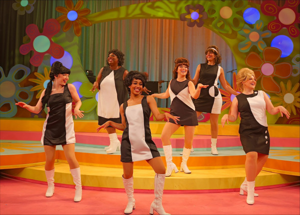  A dynamic performance moment featuring six performers in matching mod-inspired black and white mini dresses with geometric patterns and tall white go-go boots. They strike playful dance poses on a vibrant, multi-level stage decorated with oversized, colourful flower cut-outs and swirling pastel patterns. The lighting casts a warm glow on the retro set, evoking the bold style and infectious energy of the 1960s girl group era.
