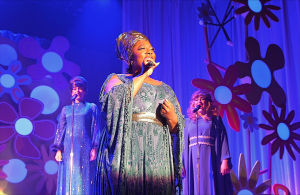 A soulful performance scene featuring a spotlighted soloist in an elegant, patterned green gown with gold accents and a matching headwrap. She holds a microphone and sings passionately downstage, while two backup singers in floor-length, shimmering blue dresses harmonize behind her. The stage is bathed in blue and purple lighting, with oversized retro flower graphics adorning the curtain backdrop, creating a rich, 1960s-inspired visual atmosphere.