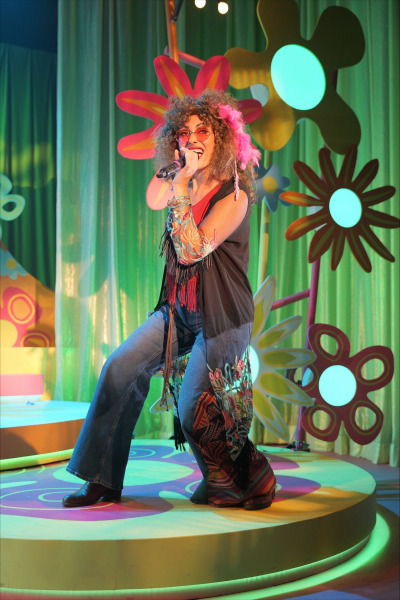 A solo performer strikes a powerful pose mid-song on a circular, psychedelic stage platform. Dressed in bell-bottom jeans with embroidered detailing, a fringed vest, and colourful feather accessories, the singer channels a 1960s hippie aesthetic. Pink-tinted sunglasses and a bold sleeve tattoo complete the vibrant look. Behind them, oversized flower power set pieces and a green glittering curtain glow under saturated theatrical lighting.