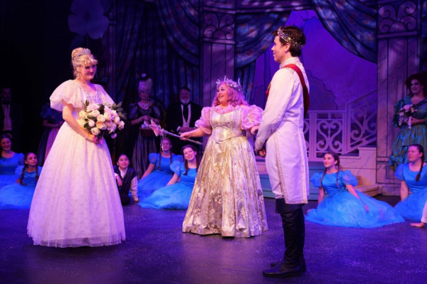 On stage in Cinderella: The Panto at King’s Wharf Theatre, a joyous royal wedding moment unfolds. Cinderella stands at left in a sparkling white ballgown and tiara, holding a bouquet of pale pink and cream roses. At centre, the Fairy Godmother — in a shimmering cream and gold gown with pastel pink puff sleeves — gestures warmly with her wand toward the Prince, who stands at right in a white royal coat with gold trim and a crown. Ensemble members dressed in bright blue costumes sit and kneel behind them, smiling and watching, with an ornate palace backdrop draped in rich blue curtains completing the magical fairy-tale tableau.