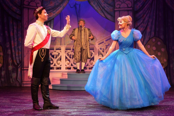  On stage in Cinderella: The Panto at King’s Wharf Theatre, the Prince stands at left in a white royal coat with gold detailing, a red sash, black boots, and a crown, raising one hand as if greeting or admiring Cinderella. At right, Cinderella wears a sparkling bright blue ballgown with puff sleeves and a glittering full tulle skirt, lifting the edges of her dress as she smiles back at him. Behind them on a small staircase, a courtier in striped period attire reacts comically, set against a whimsical palace backdrop draped in rich purple curtains.