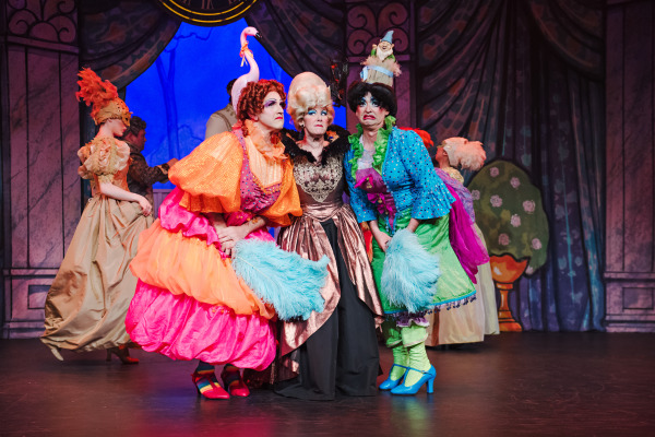 A comedic scene from a theatrical production of Cinderella: The Panto, featuring three flamboyantly dressed characters in the foreground. Two performers, portraying the stepsisters, wear exaggeratedly colourful costumes—one in pink, orange, and red with a flamingo headpiece, the other in green, blue, and purple with a garden gnome on their head. Both hold bright blue feather fans and strike dramatic expressions. Between them stands a character in a regal bronze and black gown with a tall powdered wig and a stern expression, likely the stepmother. Other masked ball attendees move about in the background against a vibrant palace backdrop.