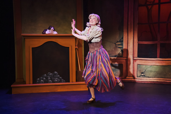 A lively moment from a theatrical production of Cinderella: The Panto, showing a character joyfully dancing in front of a painted fireplace set piece with faux coal. The performer wears a rustic costume including a striped, multi-coloured skirt, beige blouse, brown corset, and headscarf. The expression is one of happiness and freedom, with arms raised mid-dance. A small stuffed dog rests atop the fireplace, and the background is painted to resemble the interior of an old cottage.