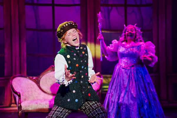 A lively moment from a theatrical production of Cinderella: The Panto, showing a performer at centre stage singing passionately, eyes closed and mouth wide open, dressed in a vest decorated with multicoloured buttons, plaid trousers, and a brown cap. Behind them stands a smiling character in a sparkling lavender gown with puffed sleeves, holding a glittery wand aloft. The scene is bathed in soft purple and pink stage lighting, with a vintage set featuring a curved-back pink loveseat and tall window panels in the background.